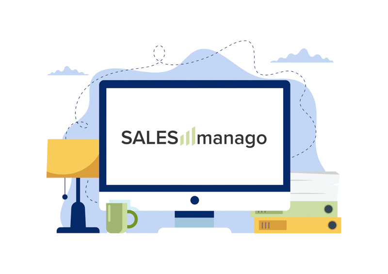 Ekran monitora z widocznym logiem sales manago. Obok stoi lampa, stos książek oraz kubek.